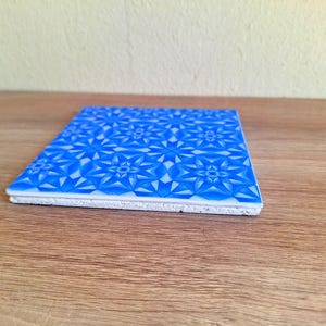 May include: A square blue and white ceramic tile with a geometric starburst pattern. The tile is resting on a wooden surface. The tile's design features a repeating pattern of blue star-like shapes against a white background.