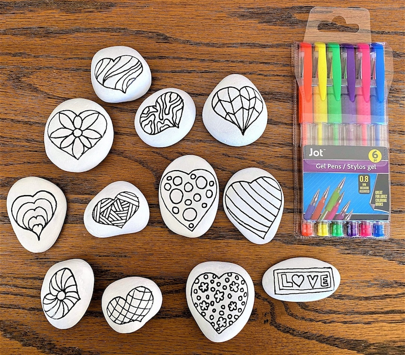 Pu&ograve; includere: Un set di 12 pietre a forma di cuore con disegni in bianco e nero. Le pietre sono disposte a semicerchio su una superficie di legno. Le pietre sono pronte per essere dipinte. Ci sono anche sei penne gel in una confezione di plastica trasparente con la parola "Jot" sul davanti.