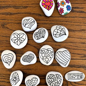 Pu&ograve; includere: Una collezione di 13 pietre dipinte a forma di cuore. Le pietre sono dipinte con vari disegni, tra cui strisce, pois, fiori e motivi geometrici. Una pietra ha la parola "LOVE" scritta su di essa.