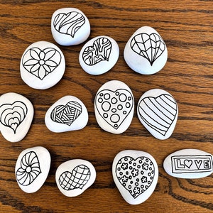 Pu&ograve; includere: Dieci pietre bianche a forma di cuore con disegni a inchiostro nero. I disegni includono un fiore, strisce, pois, spirali e la parola "LOVE".