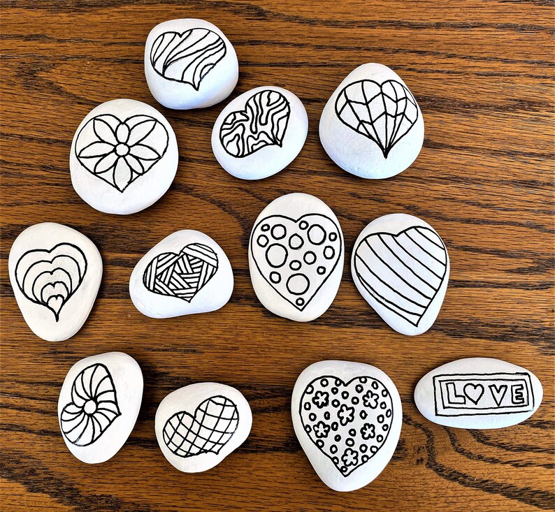 Pu&ograve; includere: Dieci pietre bianche a forma di cuore con disegni a inchiostro nero. I disegni includono un fiore, strisce, pois, spirali e la parola "LOVE".