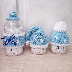 May include: Three whimsical snowman-themed decorations. The snowmen have smiling faces and are adorned with blue and white crocheted hats and accessories. One is a snow globe, and the others are marshmallow-shaped.