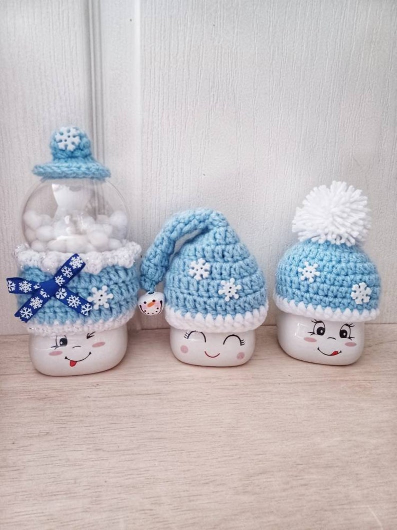 May include: Three whimsical snowman-themed decorations. The snowmen have smiling faces and are adorned with blue and white crocheted hats and accessories. One is a snow globe, and the others are marshmallow-shaped.