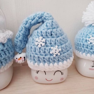 May include: A light blue crocheted hat with white snowflake accents and a small bell. The hat is on top of a white ceramic figure with a smiling face. The figure has a white base and a red smiling mouth.