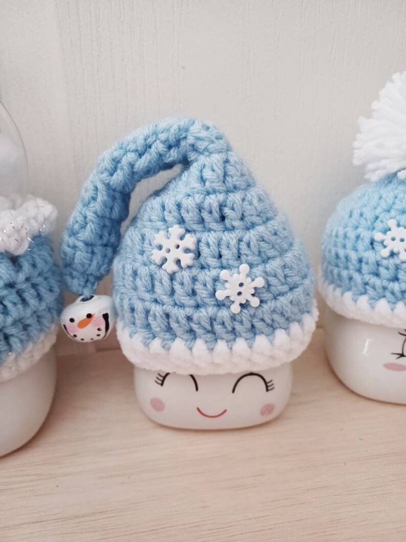 May include: A light blue crocheted hat with white snowflake accents and a small bell. The hat is on top of a white ceramic figure with a smiling face. The figure has a white base and a red smiling mouth.