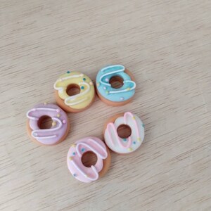 May include: Five miniature donuts with different colored glazes and sprinkles. The donuts are arranged in a circle on a light wood surface.