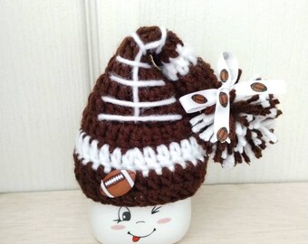 May include: A brown and white crocheted hat shaped like a football with a white pom-pom on top. The hat is decorated with a white ribbon with brown footballs printed on it. The hat is sitting on top of a white mug with a cartoon face.