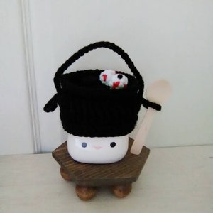 May include: A white ceramic mug with a black knitted hat and a wooden spoon. The mug has a happy face and is sitting on a wooden stand with four legs.