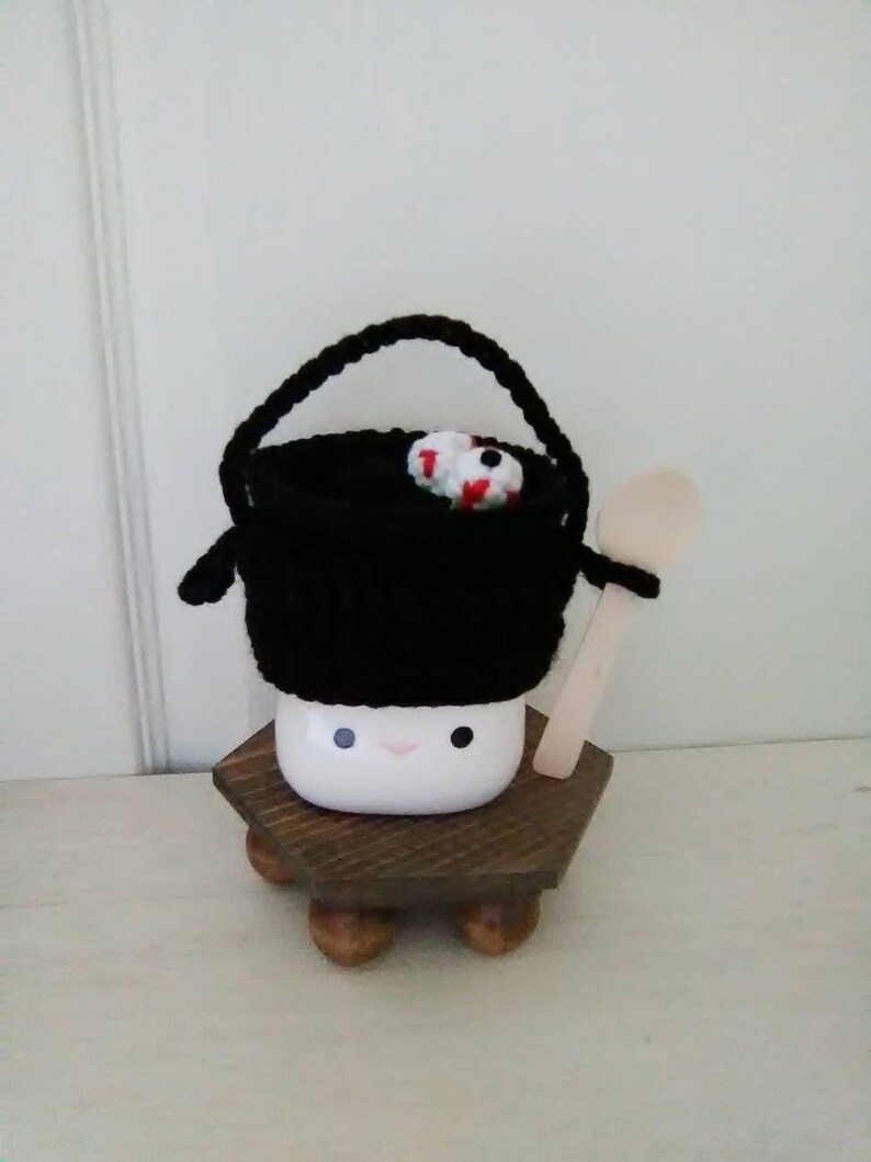 May include: A white ceramic mug with a black knitted hat and a wooden spoon. The mug has a happy face and is sitting on a wooden stand with four legs.