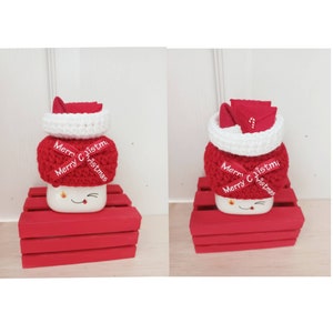 Op de afbeelding: Een witte mok met een rode gebreide kerstmanmuts en een rood lint met de tekst "Merry Christmas". De mok staat op een rode houten kist.