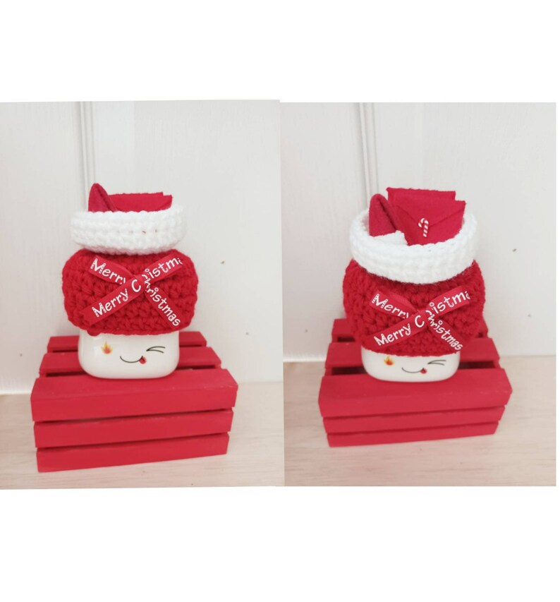 Op de afbeelding: Een witte mok met een rode gebreide kerstmanmuts en een rood lint met de tekst "Merry Christmas". De mok staat op een rode houten kist.
