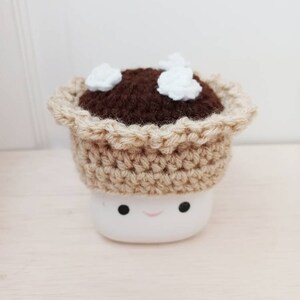 May include: A crocheted pie crust with brown filling and white marshmallow topping sits on top of a white marshmallow with a smiling face.