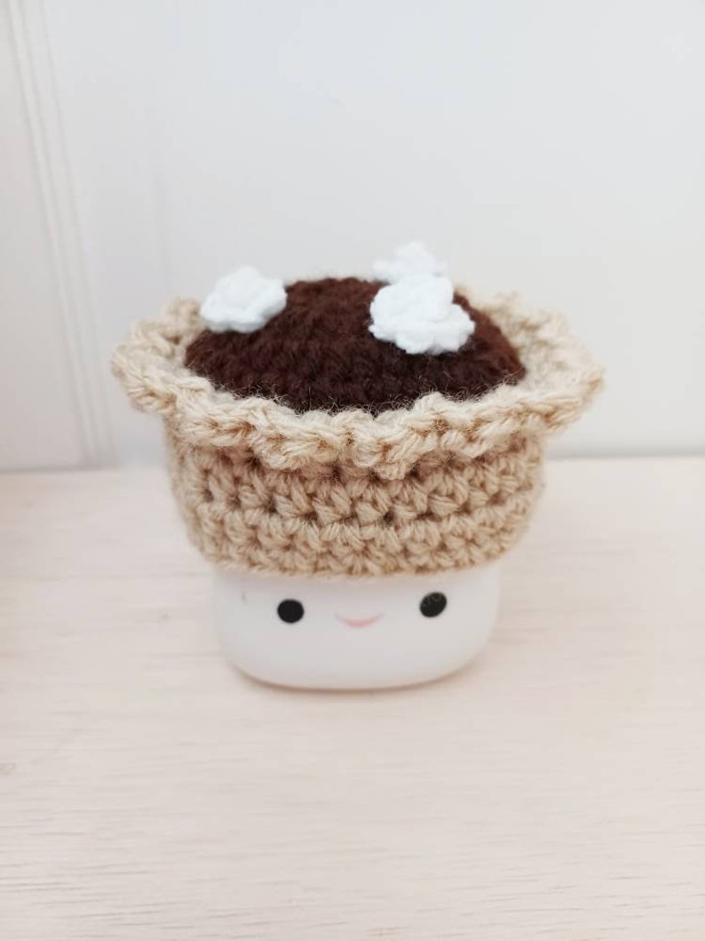 May include: A crocheted pie crust with brown filling and white marshmallow topping sits on top of a white marshmallow with a smiling face.