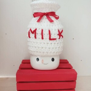 Op de afbeelding: Witte gehaakte melkpak-hoed met een rood lint en het woord "MILK" in rood geborduurd. De hoed staat op een rode houten kist.