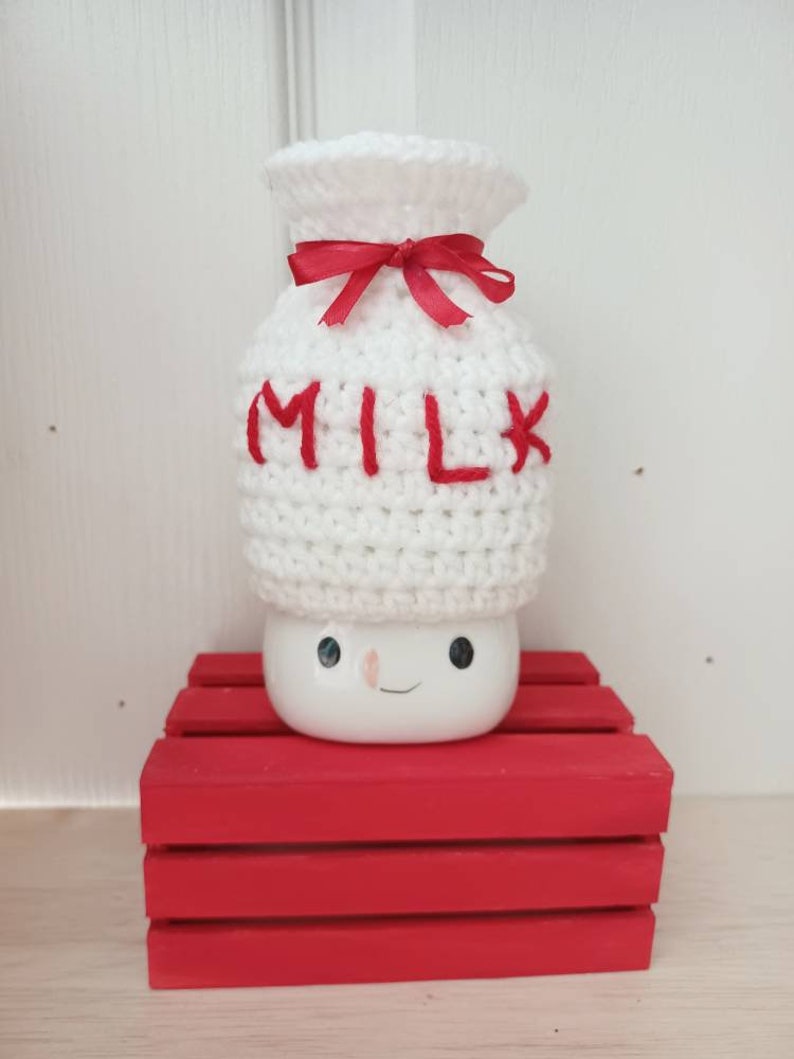 Op de afbeelding: Witte gehaakte melkpak-hoed met een rood lint en het woord "MILK" in rood geborduurd. De hoed staat op een rode houten kist.