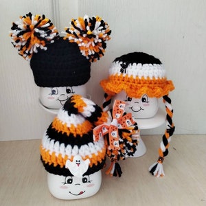 May include: Three white ceramic mugs with Halloween themed crocheted hats. The first mug has a black and orange striped hat with a ghost applique. The second mug has a black and white hat with orange trim and a black spider applique. The third mug has a black hat with orange and white pom poms.