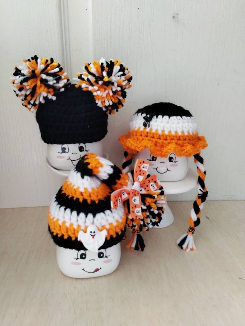 May include: Three white ceramic mugs with Halloween themed crocheted hats. The first mug has a black and orange striped hat with a ghost applique. The second mug has a black and white hat with orange trim and a black spider applique. The third mug has a black hat with orange and white pom poms.