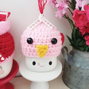 May include: A pink crocheted bird hat with black eyes and a yellow beak sits on top of a white mug with a smiling face. The mug is on a white pedestal.