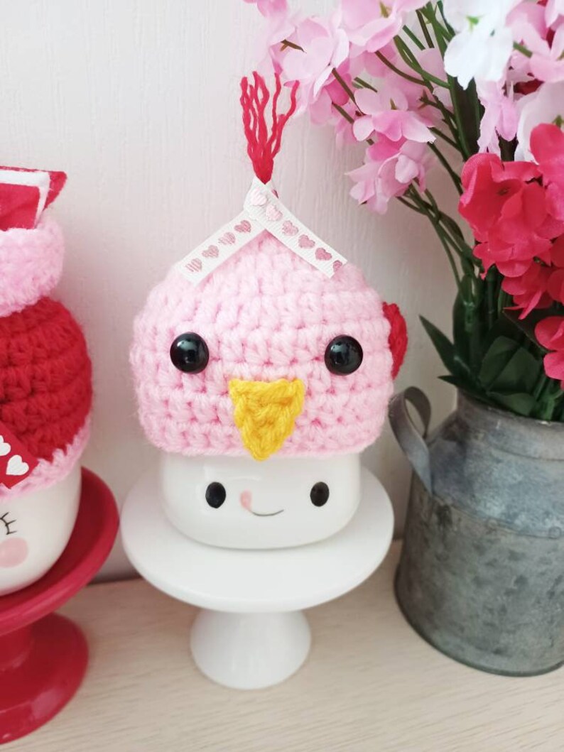 May include: A pink crocheted bird hat with black eyes and a yellow beak sits on top of a white mug with a smiling face. The mug is on a white pedestal.