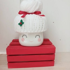 Op de afbeelding: Witte keramische mok in de vorm van een marshmallow met een witte gebreide muts met een rood lint en een groene hulstaccent. De mok heeft een glimlachend gezicht met gesloten ogen. De mok staat op een rode houten kist.