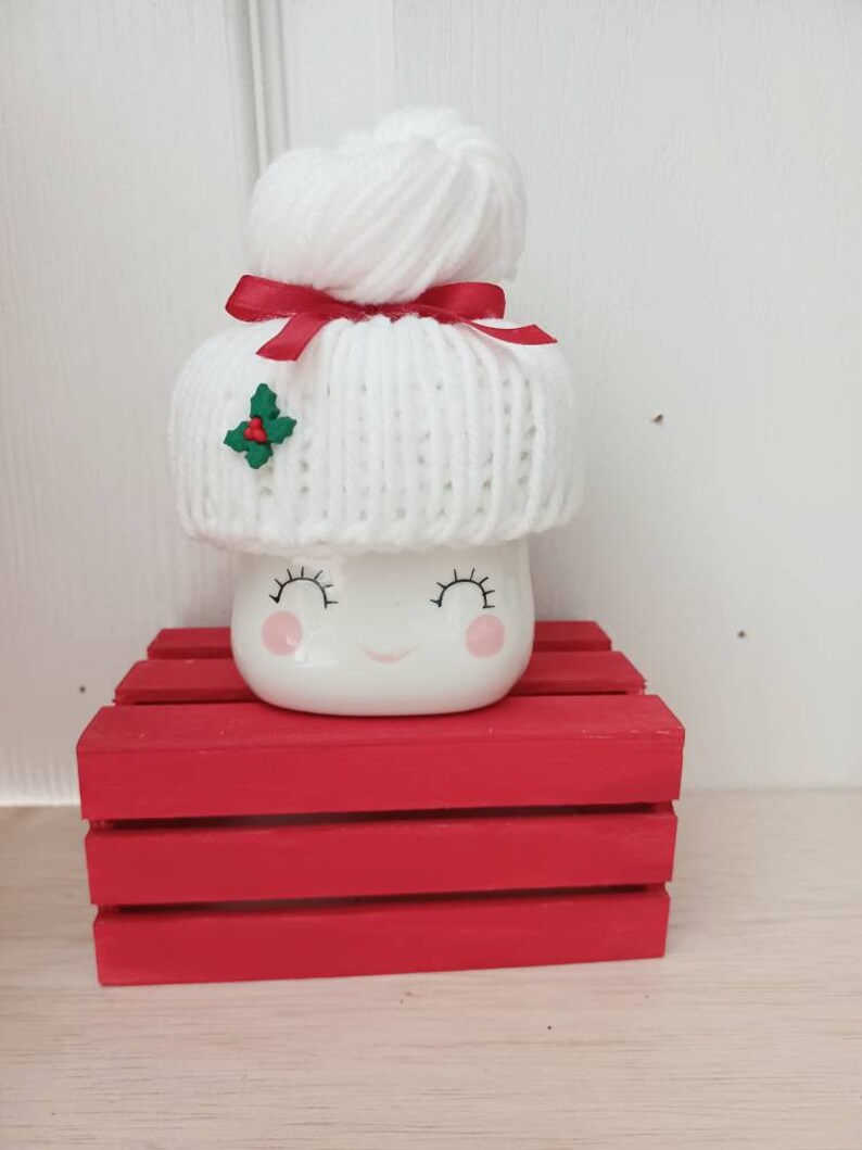 Op de afbeelding: Witte keramische mok in de vorm van een marshmallow met een witte gebreide muts met een rood lint en een groene hulstaccent. De mok heeft een glimlachend gezicht met gesloten ogen. De mok staat op een rode houten kist.