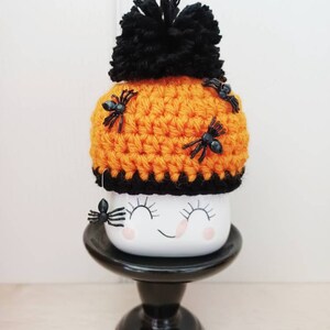 K&ouml;nnte beinhalten: Eine Halloween-Tasse mit einem l&auml;chelnden Gesicht, bedeckt mit einem orangefarbenen geh&auml;kelten Hut, der mit schwarzen Spinnen und einem schwarzen Bommel verziert ist. Die Tasse steht auf einem schwarzen Sockel und erzeugt einen gruseligen Look.