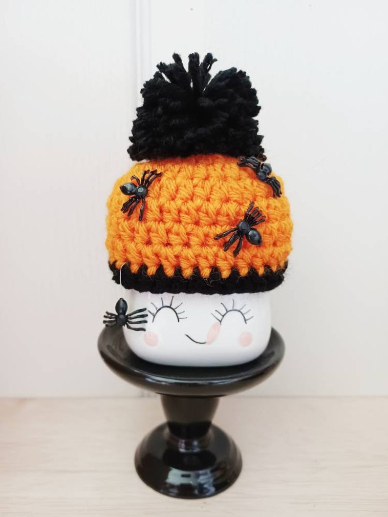 K&ouml;nnte beinhalten: Eine Halloween-Tasse mit einem l&auml;chelnden Gesicht, bedeckt mit einem orangefarbenen geh&auml;kelten Hut, der mit schwarzen Spinnen und einem schwarzen Bommel verziert ist. Die Tasse steht auf einem schwarzen Sockel und erzeugt einen gruseligen Look.