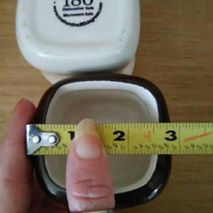 May include: Two square ceramic ramekins, one white with black lettering and one brown with a white interior. The white ramekin has the text "One Hundred 80 degrees" and "180" with a degree symbol. The ramekin is being measured with a yellow tape measure, showing a measurement of 7.6 cm.