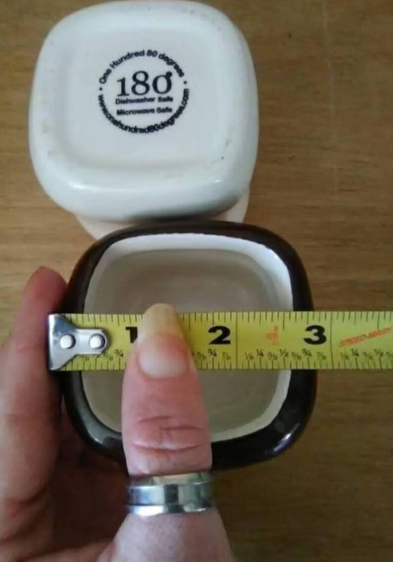 May include: Two square ceramic ramekins, one white with black lettering and one brown with a white interior. The white ramekin has the text "One Hundred 80 degrees" and "180" with a degree symbol. The ramekin is being measured with a yellow tape measure, showing a measurement of 7.6 cm.