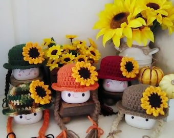 Könnte beinhalten: Eine Sammlung von Keramik-Marshmallow-Figuren, jede mit einem gehäkelten Hut und einer Sonnenblume. Die Hüte sind in verschiedenen Farben gehalten, darunter Grün, Orange und Braun, mit langen geflochtenen Haaren aus Garn. Im Hintergrund ein Strauß Sonnenblumen.