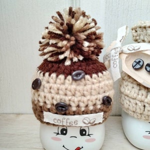 May include: Two white ceramic mugs with brown and beige crocheted hats. The hats have brown coffee bean buttons and a brown pom-pom on top. One mug has a happy face with a tongue sticking out and the text "coffee" on the hat.