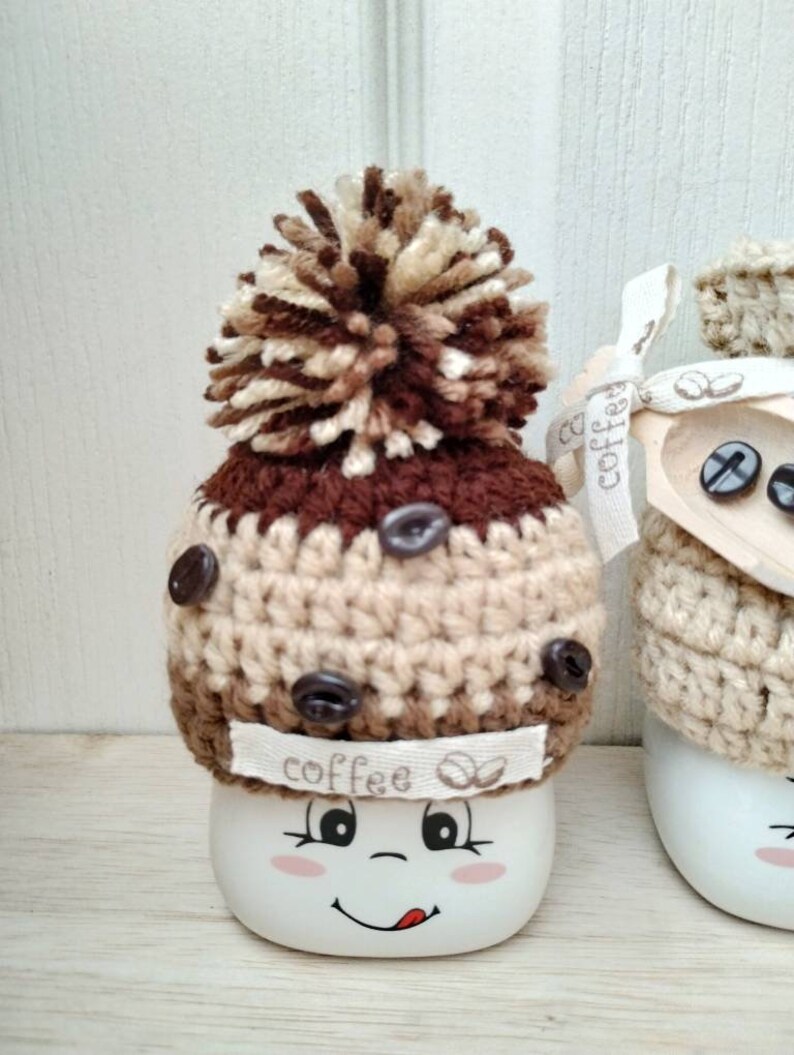 May include: Two white ceramic mugs with brown and beige crocheted hats. The hats have brown coffee bean buttons and a brown pom-pom on top. One mug has a happy face with a tongue sticking out and the text "coffee" on the hat.