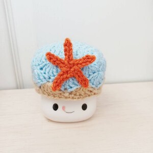 May include: A white ceramic mug with a smiling face, wearing a blue and brown crocheted hat with an orange crocheted starfish on top.