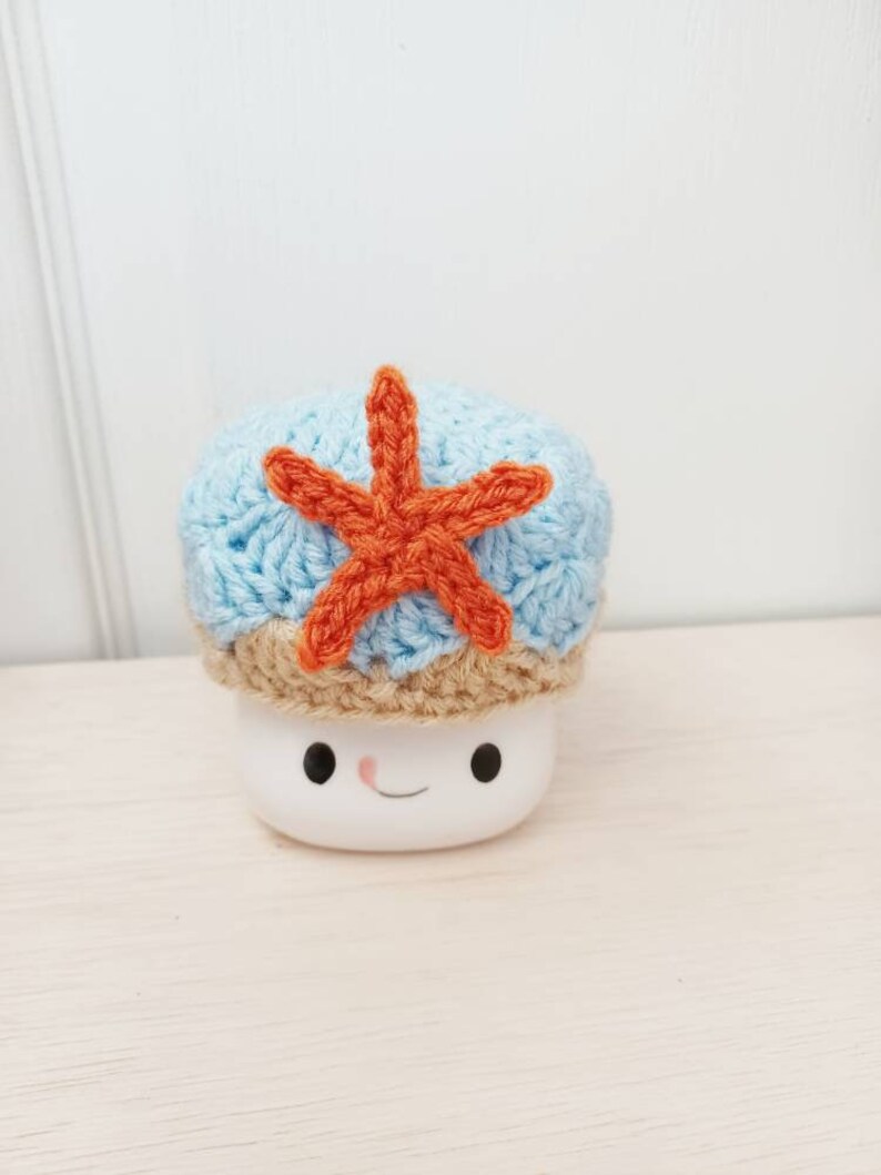 May include: A white ceramic mug with a smiling face, wearing a blue and brown crocheted hat with an orange crocheted starfish on top.