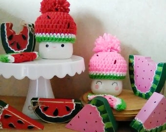Könnte beinhalten: Zwei weiße Keramikbecher mit lächelnden Gesichtern, die gehäkelte Wassermelonenhüte tragen. Die Hüte sind rot und pink mit grünen Akzenten. Es gibt auch mehrere bemalte Wassermelonenstücke aus Holz in Rot, Pink und Grün.