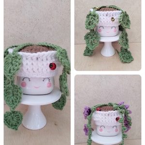 May include: Three white ceramic mugs with smiling faces are decorated with crocheted hats and greenery. The mugs are on white cake stands. The hats are white with brown yarn and the greenery is green and purple.