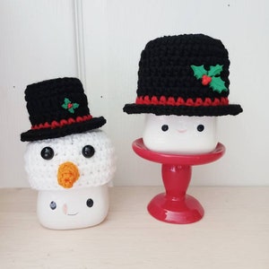 May include: Two white ceramic mugs with snowman faces, one wearing a black crocheted hat with a red trim and a green holly accent, the other wearing a black crocheted hat with a red trim and a green holly accent, sitting on a red pedestal.