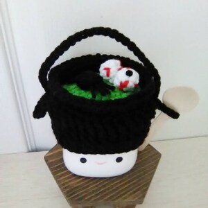 May include: A black crocheted cauldron with a green lining and two googly eyes sits on a wooden stand with a smiling face.