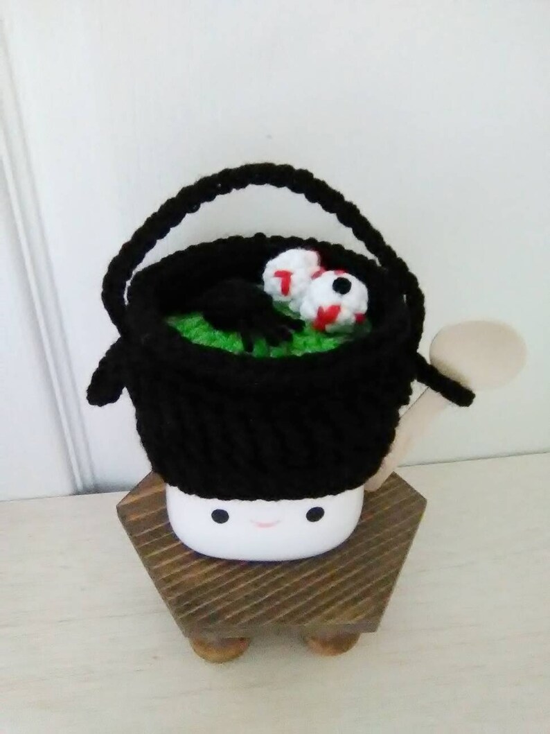 May include: A black crocheted cauldron with a green lining and two googly eyes sits on a wooden stand with a smiling face.