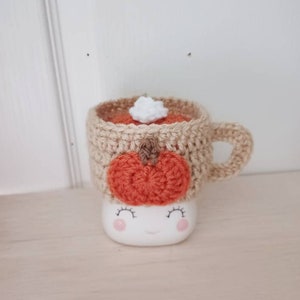 May include: A crocheted mug cozy with a pumpkin design and a white crocheted marshmallow on top. The cozy is beige and the pumpkin is orange. The mug cozy is on a white mug with a smiling face.