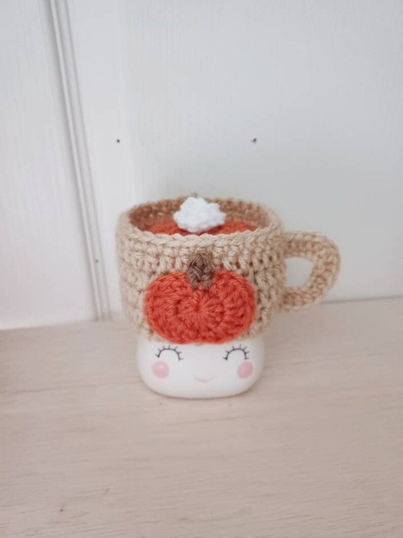 May include: A crocheted mug cozy with a pumpkin design and a white crocheted marshmallow on top. The cozy is beige and the pumpkin is orange. The mug cozy is on a white mug with a smiling face.