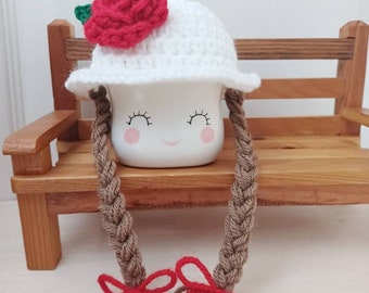 May include: A white ceramic mug with a smiling face, wearing a white crocheted hat with a red rose on top and brown yarn braids with red bows.