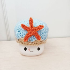 May include: A white ceramic mug with a smiling face, wearing a blue and brown crocheted hat with an orange crocheted starfish on top.