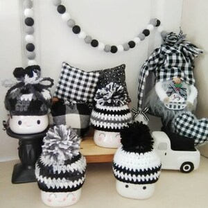May include: Five white ceramic mugs with black and white crocheted hats. The mugs have a smiling face and pink cheeks. The hats have pom-poms on top. There is a black and white gnome and a white toy truck in the background.