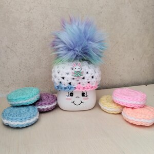 May include: A white mug with a smiling face, topped with a crocheted hat with a rainbow pom-pom and a bunny charm. The mug is surrounded by crocheted macarons in various pastel colors.