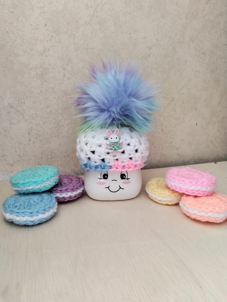 May include: A white mug with a smiling face, topped with a crocheted hat with a rainbow pom-pom and a bunny charm. The mug is surrounded by crocheted macarons in various pastel colors.