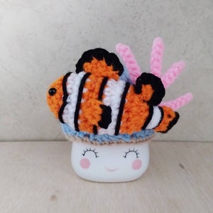 May include: A crocheted clownfish with orange, white and black stripes sits on top of a white mug with a smiling face. The clownfish has a pink coral attached to it.