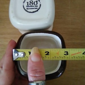 May include: Two square ceramic ramekins, one white with a blue logo that says "One Hundred 80 degrees" and "180" and the other black with a white interior. A yellow measuring tape is wrapped around the black ramekin, measuring 3 inches in diameter.