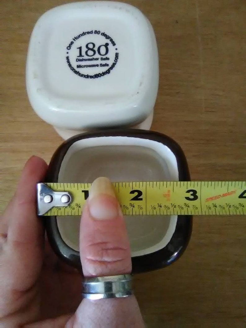 May include: Two square ceramic ramekins, one white with a blue logo that says "One Hundred 80 degrees" and "180" and the other black with a white interior. A yellow measuring tape is wrapped around the black ramekin, measuring 3 inches in diameter.