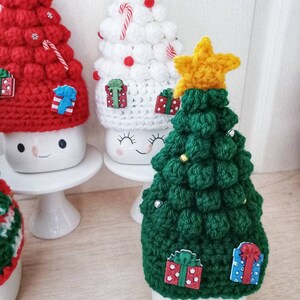May include: Three crocheted Christmas tree hats in red, white, and green. Each hat has a star on top and is decorated with candy canes, presents, and beads. The hats are sitting on top of white mugs with cute faces.